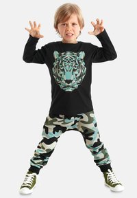 Schwarzes Langarmshirt mit einem geometrischen Tiger-Graphic, kombiniert mit beigefarbenen, grünen und schwarzen Camouflage-Sweatpants. Weiße Sneaker.