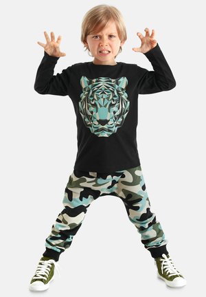 Schwarzes Langarmshirt mit einem geometrischen Tiger-Graphic, kombiniert mit beigefarbenen, grünen und schwarzen Camouflage-Sweatpants. Weiße Sneaker.