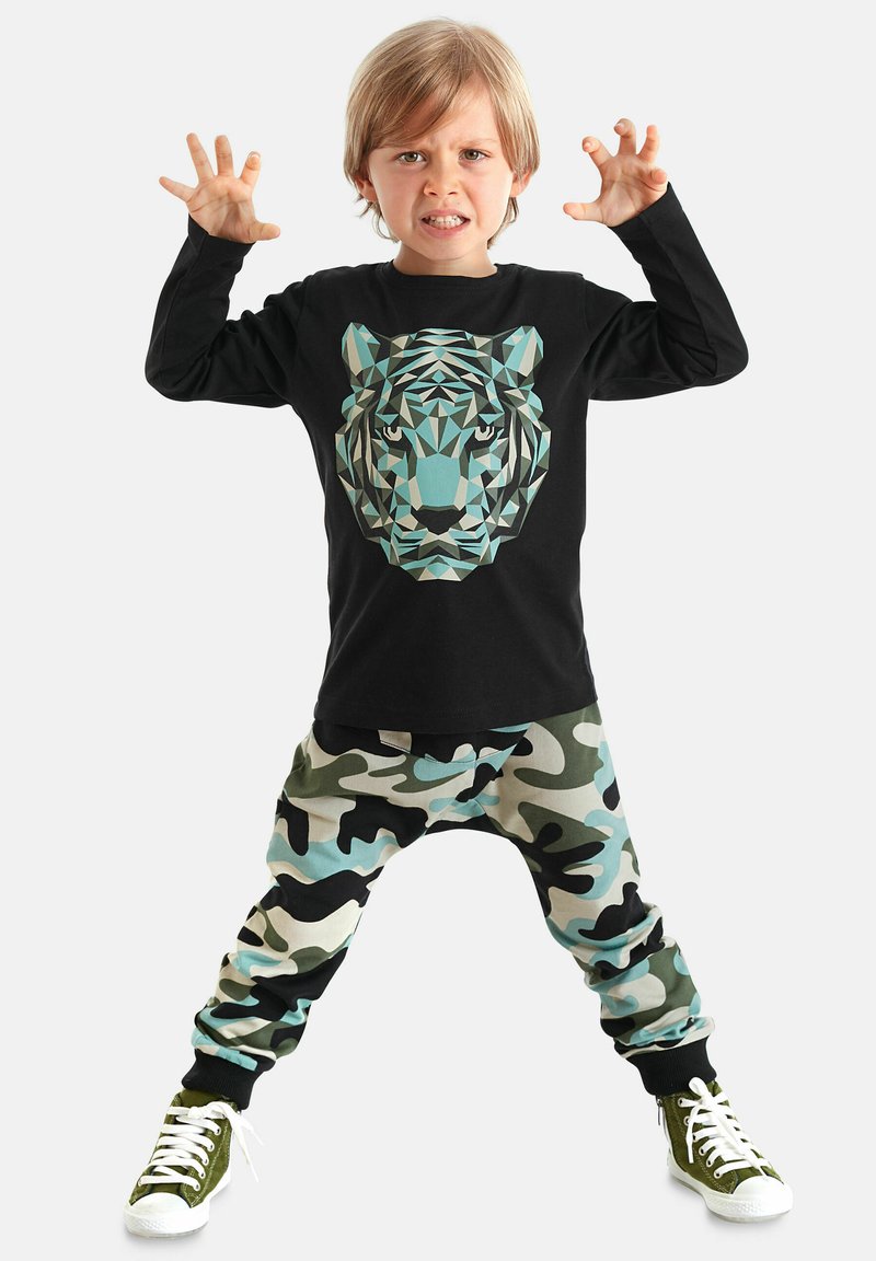Schwarzes Langarmshirt mit einem geometrischen Tiger-Graphic, kombiniert mit beigefarbenen, grünen und schwarzen Camouflage-Sweatpants. Weiße Sneaker.