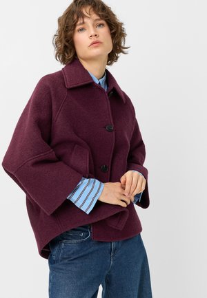Frau trägt einen burgunderfarbenen Wollmantel über einem blau gestreiften Hemd und blauen Jeans, steht mit gefalteten Händen vor einem schlichten Hintergrund.