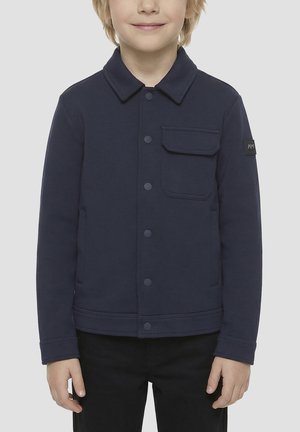 Enfant portant une veste bleu marine boutonnée avec une poche poitrine et un pantalon noir sur un fond clair uni.