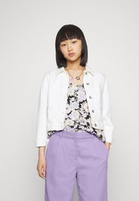 Jaqueta de ganga branca, top preto com flores e calças lilás. Colar de corrente dourada com detalhes roxos. Texturas suaves e corte descontraído.