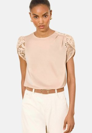 Kobieta nosząca beżową bluzkę z krótkimi koronkowymi rękawami, w połączeniu z białymi spodniami z wysokim stanem i brązowym paskiem ze złotą klamrą.