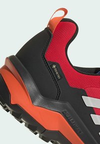 Punainen ja musta vaelluskengä, jossa on verkkonetti yläosa, kuvioitu kumipohja, oranssit yksityiskohdat ja GORE-TEX -merkki vedenpitävää suojaa varten.