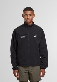 Giovane uomo con una giacca nera con zip e loghi bianchi e pantaloni corti verde oliva, in piedi su uno sfondo grigio chiaro uniforme.