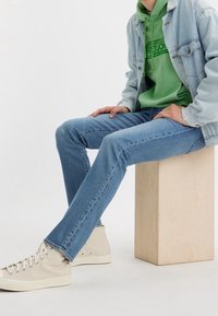 Personne assise sur un bloc de bois portant un jean bleu clair, des baskets montantes beiges, un sweat à capuche vert et une veste en jean claire.