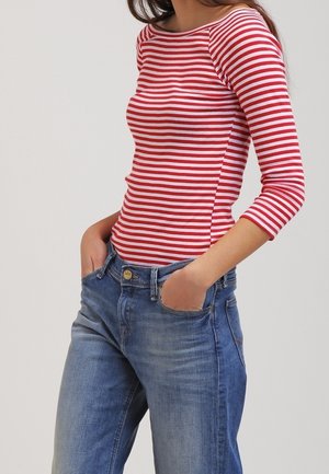 Frau trägt rot-weiß gestreiftes Langarmshirt und blaue Jeans, steht mit Händen in den Taschen vor einfachem Hintergrund.