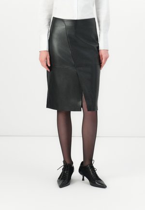 Kvinne iført et svart lær skjørt til knærne med en frontslitz, svarte gjennomsiktige strømpebukser, svarte snørel boots med hæl og en hvit langermet skjorte.