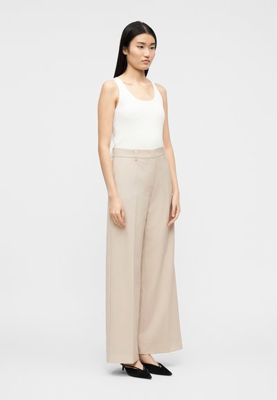 Γυναίκα που στέκεται με ίσια μαύρα μαλλιά, φοράει λευκό μπλουζάκι χωρίς μανίκια, μπεζ π αντελόνι φαρδύ στο πόδι και μαύρες μυτερές γόβες, πάνω σε απλό φόντο.