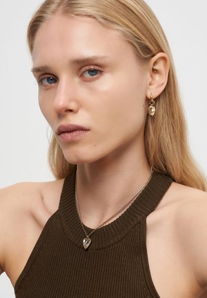 Gul hjerteformede øreringe og en sølv hjertevedhæng halskæde båret over en mørk ribbet ærmeløs top. Glatte teksturer og rene linjer.