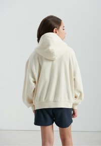 Sweat à capuche en polaire de couleur blanc cassé, doté d'une capuche, d'une coupe oversize, de poignets élastiques et d'une surface texturée, associé à un short bleu marine.