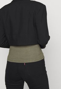 Blazer noir court à manches longues porté sur un pull côtelé vert olive ; pantalon noir avec un onglet rouge visible sur la poche arrière.