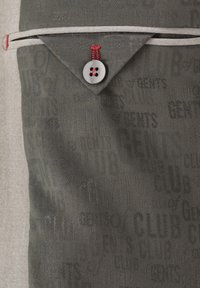 Nahaufnahme einer grauen Stofftasche mit schwarzen Textmustern, einer Klappe, die durch einen grauen Knopf mit rotem Faden gesichert ist, und weißem Besatz auf beigem Hintergrund.