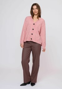 Roze gebreide cardigan met V-hals en zwarte knopen, gecombineerd met bruine geruite broek en puntige zwarte schoenen. Losse pasvorm en zachte textuur.