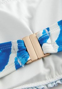 Witte zwemkledingstof met een blauwe tie-dye accent en een goudkleurige rechthoekige sluiting, met een gestructureerde afwerking en een gerimpelde zoom.