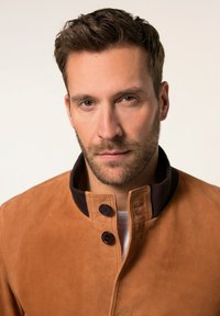 Lichtbruine suède jas met een staande kraag, voorzien van twee donkerbruine knopen en een gladde textuur. Effen wit shirt eronder.
