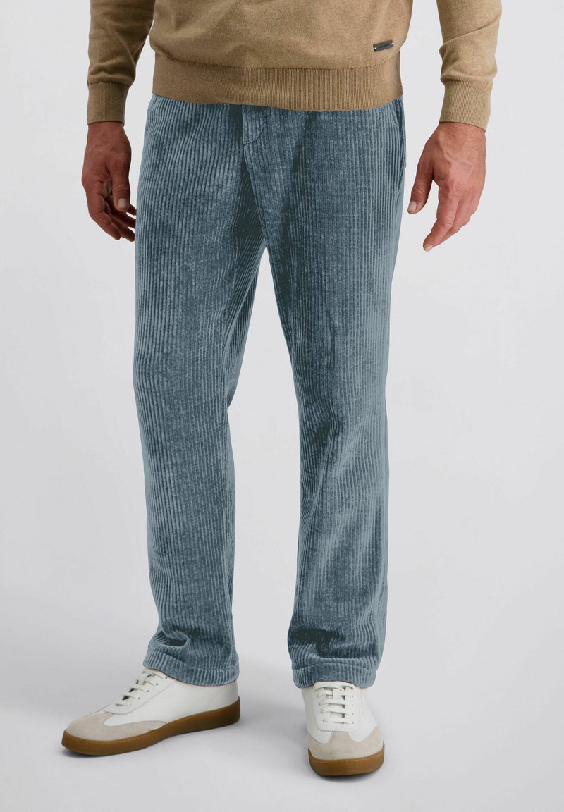 Man in een lichtblauwe corduroy broek, beige trui en witte sneakers met bruine zolen, staande tegen een effen achtergrond.