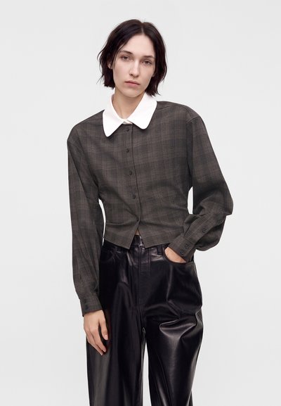 Jeune femme aux cheveux courts et foncés porte un chemisier à carreaux foncé boutonné avec un col blanc et un pantalon large en cuir noir, debout devant un fond blanc.