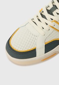 Sneaker met een crèmekleurige perforeerde leren bovenkant, groene accenten en gele details. Voorzien van platte veters en een gestructureerde zool.