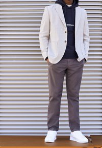 Ljusgrå rutigt kavaj över en mörk huvtröja, matchad med grå byxor och vita sneakers. Kavajen har två knappar och fackfickor.