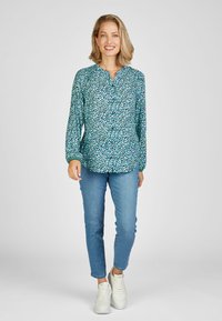 Helles blaues, gemustertes Oberteil mit langen Ärmeln, V-Ausschnitt, kombiniert mit hellblauen Jeans und weißen Sneakern. Einfaches, lässiges Outfit.