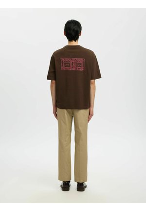 Personne aux cheveux courts et foncés vue de dos, portant un t-shirt marron avec un motif de devanture rouge et un pantalon beige, sur un fond clair uni.