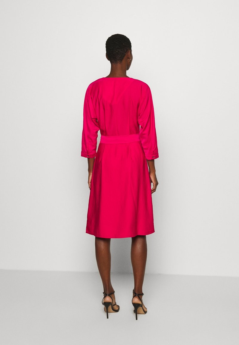 Femme portant une robe rose vif arrivant au genou avec des manches trois-quarts et des sandales à talons noirs, se tenant devant un fond blanc uni.