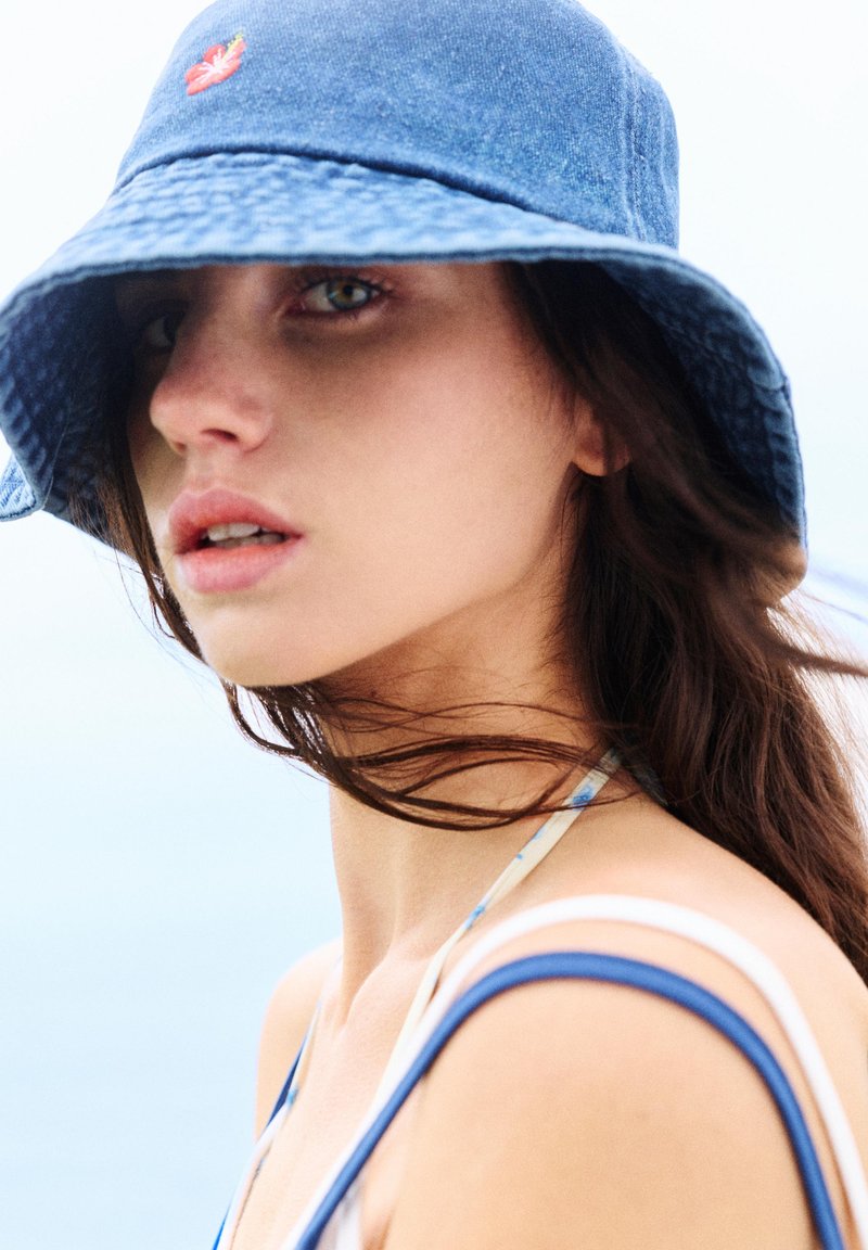 Jeune femme aux longs cheveux foncés porte un chapeau bob bleu et un top à fines bretelles, regardant de côté sur un fond de ciel pâle.