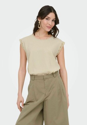 Femme aux cheveux foncés portant un haut beige sans manches avec une bordure en dentelle et un pantalon ample taille haute kaki sur un fond uni.
