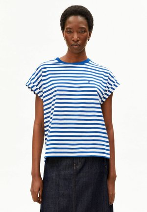 Femme aux cheveux courts et bouclés portant un t-shirt à manches courtes rayé bleu et blanc et une jupe en denim foncé, debout devant un fond blanc uni.