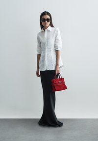 Camicia bianca con bottoni e dettagli testurizzati, gonna nera lunga fino a terra e una piccola borsa in suede rossa con manico superiore. Occhiali da sole neri.