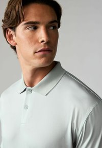 Hellgraues Poloshirt mit zwei Knöpfen, einem gerippten Kragen und einem subtilen strukturierten Muster. Der Stoff wirkt glatt und leicht.