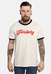 Camiseta de algodón blanca con ribete negro y texto "Forsberg" en rojo. Mangas cortas, cuello redondo, que presenta brazos tatuados y jeans de mezclilla.