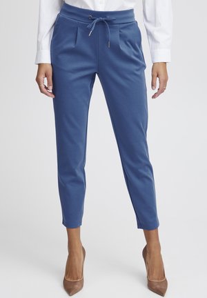 Frau trägt blaue, schmal zulaufende Hose mit Kordelzug in der Taille, weißes langärmeliges Hemd und braune spitze Pumps, steht vor neutralem Hintergrund.