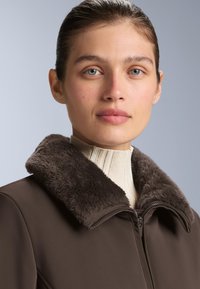 Hnedý kabát s jemným umelým kožúškom, so zipsovým uzáverom a rebrovaným krémovým rolákom pod ním. Hladká textúra látky.