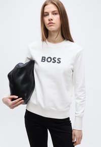 Νεαρή γυναίκα με λευκό φούτερ που αναγράφει "BOSS", κρατώντας μια μεγάλη μαύρη τσάντα φάκελο, πάνω σε απλό φωτεινό φόντο.