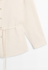 Chemise crème à manches longues avec boutons, ceinture ajustable à la taille et ourlet droit, présentée sur fond blanc.
