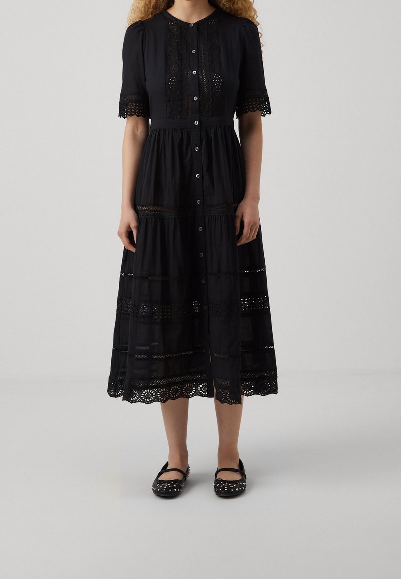 Robe midi noire avec fermeture à boutons sur le devant, détails en dentelle sur les manches et la jupe, portée avec des chaussures Mary Jane noires cloutées.