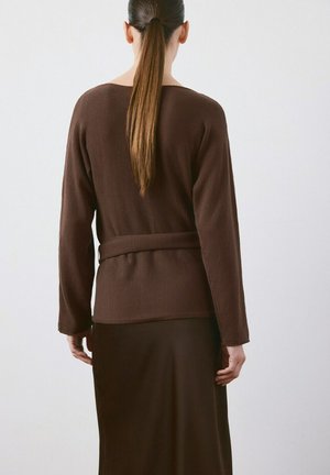 Femme avec une longue queue de cheval portant un pull marron ceinturé et une jupe assortie, debout face à un mur blanc uni.