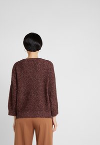 Pull tricot oversize bordeaux avec motif texturé, poignets et ourlet côtelés. Le modèle porte un pantalon beige à jambes larges.