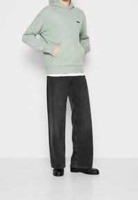 Sudadera con capucha de color verde claro y bolsillo frontal, superpuesta sobre una camisa blanca, combinada con jeans amplios oscuros y zapatos negros.