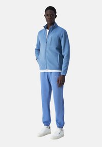 Giacca a zip in fleece azzurro chiaro con una tasca, abbinata a pantaloni jogger coordinati. Sneakers bianche completano l'outfit. Tessuto morbido, design casual.