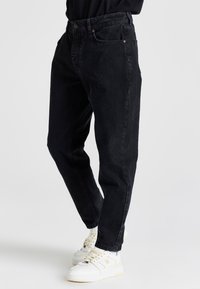 Jeans de mezclilla negra con un corte relajado, que cuentan con cinco bolsillos, detalles de costura sutiles y una cintura con cierre de botón, llevados con zapatillas blancas.