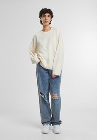 Oversized white sweater with ribbed cuffs and hem paired with light blue distressed jeans featuring large knee rips and white sneakers.