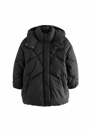 Personne portant une doudoune noire avec poignets et ourlet élastiques, debout à l'intérieur devant un fond en bois et blanc.