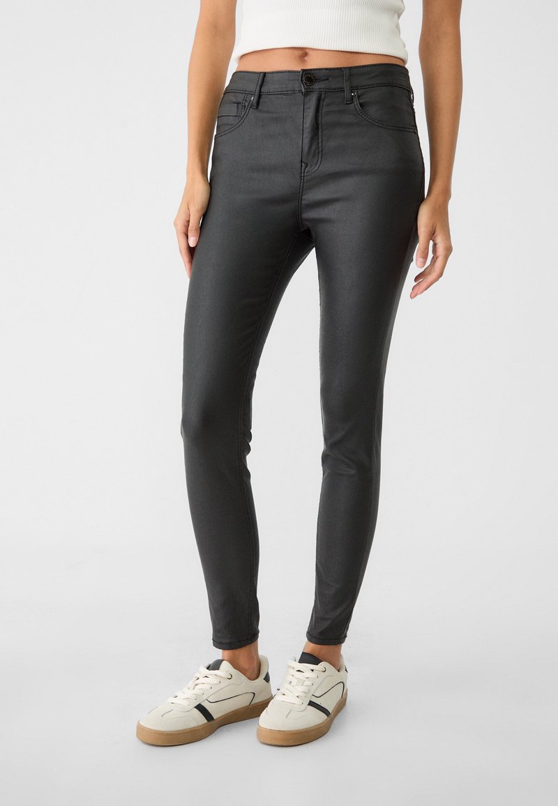 Črne skinny hlače iz jeansa z gladko, sijajno teksturo, zasnovo s petimi žepi in zapenjanjem na gumb, v kombinaciji z belimi supergami z temnimi detajli.