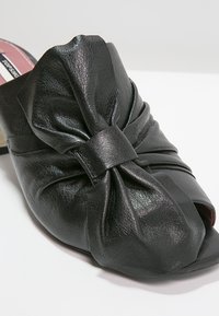 Mule en cuir noir avec un grand nœud torsadé sur le dessus et un bout ouvert, présenté sur fond blanc.