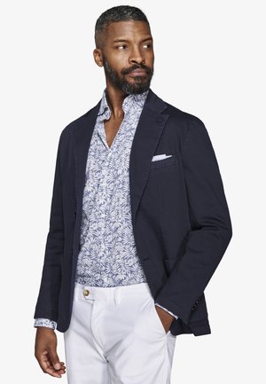 Mężczyzna w granatowej marynarce na niebiesko-białej koszuli w kwiaty i białych spodniach, patrzący w lewo, z jedną ręką w kieszeni.