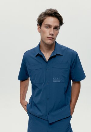 Mladý muž so svetlohnedými vlasmi, oblečený v modrej košeli s krátkym rukávom a nohaviciach rovnakej farby, stojaci s rukami v bodoch na jednoduchom pozadí.