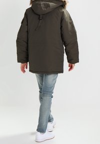 Olijfgroene parka met een met bont gevoerde capuchon, geborduurde textuur en verstelbare koord. Gecombineerd met lichtblauwe jeans en witte sneakers.